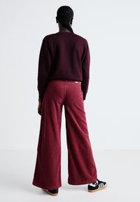 Pantaloni largi din catifea în culoarea vișiniu, cu o suprafață texturată, asortați cu un hanorac vișiniu și adidași negri cu dungi albe.