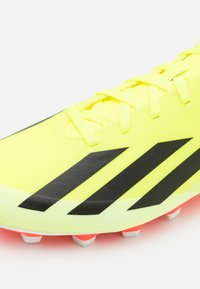 Botín de fútbol de color amarillo brillante con rayas diagonales negras, parte superior sintética texturizada, con cordones en la parte delantera y acentos en blanco y naranja en las suelas.