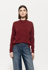 Frau mit glattem braunem Haar, die goldene Creolen trägt, einen bordeauxfarbenen Langarmpullover sowie hochtaillierte blaue Jeans mit vorderen Taschen.