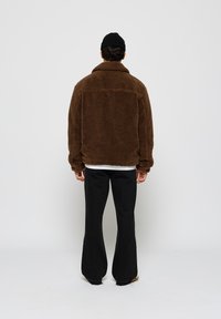 Brun fleecejakke med høy krage, oversized passform og teksturert overflate, kombinert med svarte utsvingte bukser, sett bakfra.