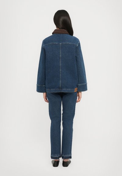 Oseba stoji obrnjena stran, oblečena v temno modro jeans jakno z rjavim ovratnikom, ujemajoče se ravne jeans hlače in črne čevlje.