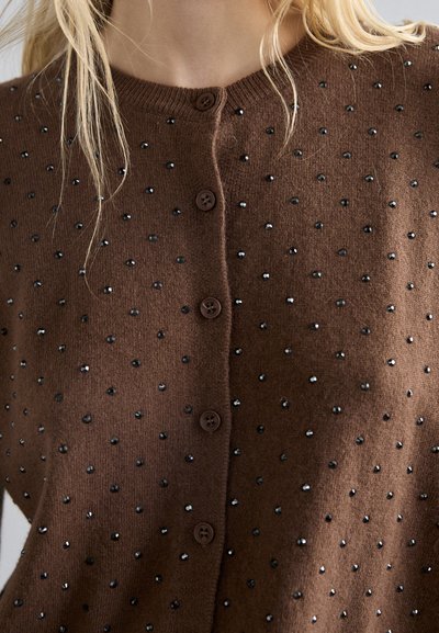 Καφέ πλεκτό καρντιγκάν με στρογγυλά κουμπιά και διασκορπισμένες μαύρες χάντρες, που επιδεικνύει ένα υφασμάτινο σχέδιο με υφή και απαλότητα.
