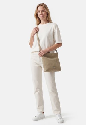 Naine, kes kannab valget t-särki, helebeežeid pükse ja valgeid tenniseid, hoides käes beeži värvi õlakoti rihmaga üheltaustal.