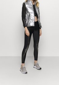 Silverfärgad vadderad väst med två framfickor, kombinerad med svarta leggings som har en matt och glänsande textur, samt ljusgrå träningsskor.