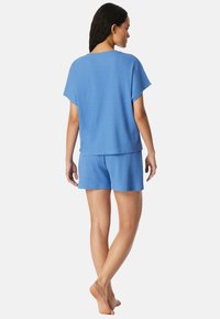 Frau steht barfuß, mit dem Rücken zur Kamera, trägt ein locker sitzendes blaues Kurzarmoberteil und passende Shorts vor einem einfarbigen weißen Hintergrund.