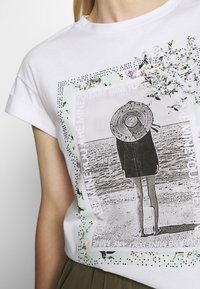 Camiseta de algodón blanca con un gráfico en blanco y negro de una figura junto al agua, rodeada de acentos florales y texto en colores pastel.