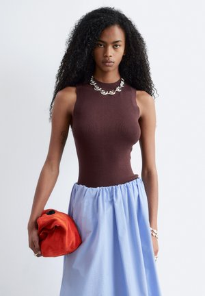 Femme aux longs cheveux bouclés, portant un haut marron sans manches et une jupe bleu clair, tenant une pochette rouge, accessoirisée d'un collier et d'un bracelet en argent.