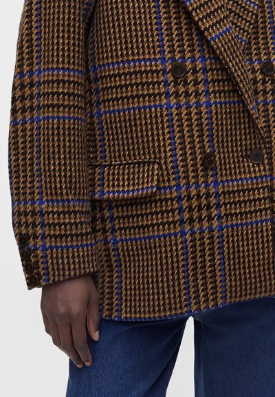 Καφέ και μαύρο σακάκι με σχέδιο houndstooth και μπλε μοτίβο παραθύρου, μαύρα κουμπιά, πλαϊνή τσέπη με καπάκι, φορεμένο με σκούρα μπλε τζιν σε σκουρόχρωμο χέρι.