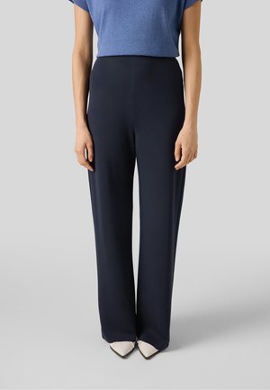 Kobieta ubrana w granatowe, szerokie spodnie z wysokim stanem, niebieską bluzkę z krótkim rękawem oraz białe buty z noskiem w szpic, stoi na jednolitym jasnym tle.