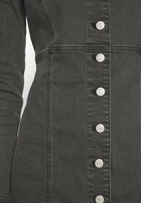 Gros plan sur le devant d'une veste en denim gris foncé avec une rangée verticale de boutons-pression argentés et des détails de couture visibles.