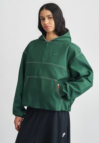 Veste à capuche verte avec détails de couture blancs, fermeture éclair à l'avant et poches latérales, associée à un pantalon noir avec un logo.