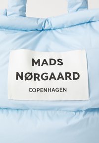 Ljust blå pufferjacka med en framträdande vit etikett med svart text som säger "MADS NØRGAARD COPENHAGEN." Slät nylonyta.