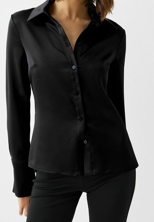 Camicia nera in satin con colletto, maniche lunghe e una texture liscia. Presenta un orlo curvo e sottili accenti di bottoni.