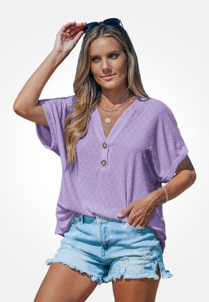 Vrouw die haar zwarte zonnebril rechtzet, gekleed in een lavendelkleurige blouse met korte mouwen en ooglets, met knopen, en lichtblauwe beschadigde denim shorts.