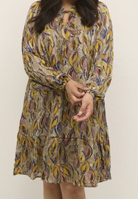 Robe à manches longues avec un motif de feuilles colorées en jaune, rouge et bleu sur un fond beige, dotée d'une encolure avec ouverture en forme de goutte et de poignets froncés.