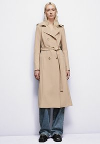 Béžový trenčkot s dvojitým zapínaním, gombíkovými detailmi a zaviazaným opaskom, doplnený o široké modré džínsy a čierne topánky.