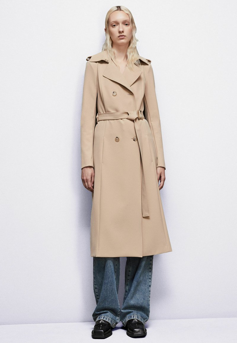 Béžový trenčkot s dvojitým zapínaním, gombíkovými detailmi a zaviazaným opaskom, doplnený o široké modré džínsy a čierne topánky.