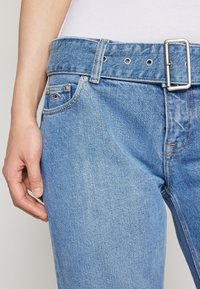 Vaqueros de mezclilla de color azul claro con un lazo de cinturón ancho, hebilla rectangular de plata y un bolsillo pequeño que presenta un emblema rojo y azul.