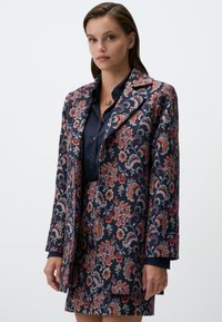 Blazer navy à motif floral avec un design paisley multicolore, coupe ajustée, revers cranté et deux poches avant, porté sur un haut sombre.