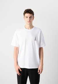 Weißes Baumwoll-T-Shirt mit kurzen Ärmeln, Rundhalsausschnitt und einem kleinen schwarzen Logo auf der linken Brustseite. Einfaches Design, glatte Textur.