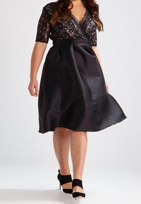 Svart klänning med spetsbodice, korta ärmar och en satin kjol. Har en V-urringning och en figurnära midja, som flödar till längden mitt på vaden.