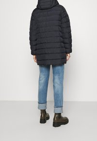 Osoba w czarnej pikowanej kurtce, z wywiniętymi jasnoniebieskimi dżinsami i ciemnooliwkowych, masywnych botkach za kostkę, stojąca na tle gładkiej białej ściany.