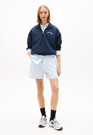 Kobieta stojąca, ubrana w granatową bluzę z kołnierzem na suwak, jasnoniebieską, pasiastą koszulę i krótkie spodenki, czarne skarpetki oraz srebrne trampki na białym tle.