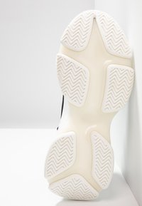 Suela de zapatilla deportiva blanca con un patrón de banda de rodadura en zigzag, que presenta un caucho liso de color claro con secciones angulares y un detalle de acento negro.