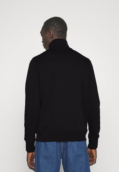 Sweatshirt noir à col roulé avec des manches longues, des poignets côtelés et un ourlet. Texture lisse, design ajusté, porté avec un pantalon en denim bleu.