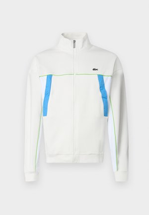 Bela zip-up jakna z modrimi in zelenimi poudarki. Ima visok ovratnik, stranske žepe in logo na prsih. Gladka struktura tkanine.