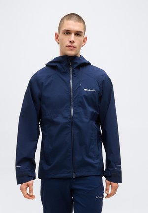 Hombre vestido con chaqueta y pantalones azul marino de Columbia, parado frente a un fondo claro y liso, mirando al frente con expresión neutral.