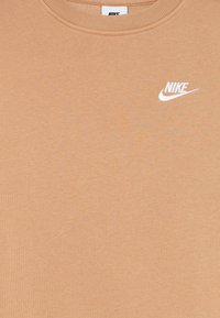 Beige Nike póló fehér hímzett logóval a bal felső mellen és kerek nyakkivágással.