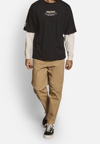 Sort T-shirt med korte ærmer med teksten "GRACIOUS", lagdelt over en lys langærmet skjorte, parret med beige chinos og sorte sneakers.