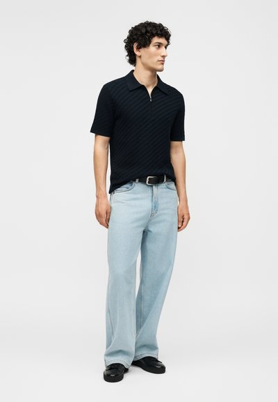 Άνδρας με σγουρά μαλλιά που φοράει μαύρο κοντομάνικο πόλο με υφή, ανοιχτό μπλε φαρδιές τζιν, μαύρη ζώνη και μαύρα παπούτσια, στέκεται και κοιτάζει στο πλάι.