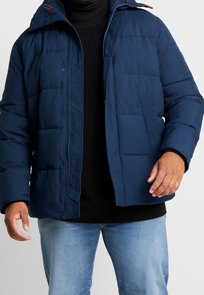 Marineblå dynejakke med lynlås foran, trykknapper og quiltet tekstur. Har hætte og sidelommer. Båret over en sort sweater.