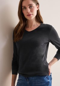 Top nero a maniche lunghe con scollo a V, realizzato in morbido tessuto e con polsini a coste, abbinato a jeans in denim blu. Il modello è in piedi di fronte a un muro chiaro.