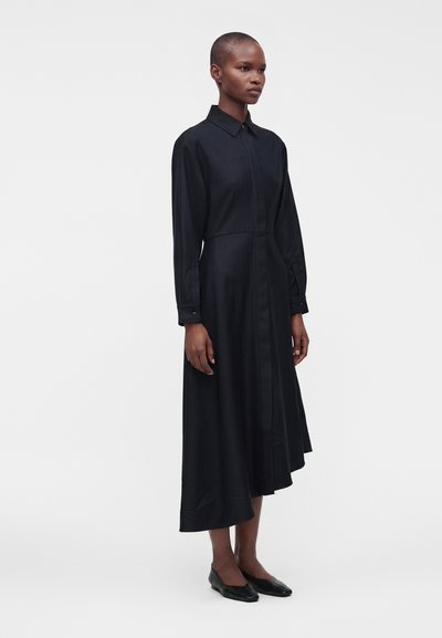 Femme debout portant une robe chemise midi noire à manches longues avec col et des ballerines noires sur un fond blanc uni.