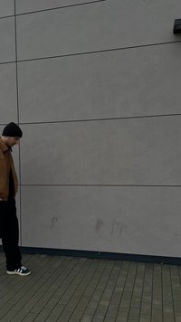 Giacca di pelle marrone, camicia nera, pantaloni neri e berretto nero indossati da una persona in piedi su una superficie lastricata accanto a una parete grigia testurizzata.