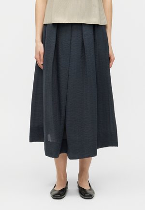 Vrouw draagt een donkerblauwe, geplooide rok tot halverwege de kuit, gecombineerd met zwarte platte schoenen en een beige mouwloos topje.