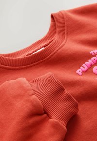 Primer plano del puño de una sudadera rojo-naranja y el cuello acanalado con texto parcial rosa que dice ""PUMPK"" visible en la tela.