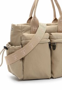 Beiger, gepolsterter Tragetasche mit zwei vorderen Reißverschlusstaschen, verstellbarem, markengeprägtem Stoffriemen, kurzen Griffen und seitlicher elastischer Kordelzugtasche.