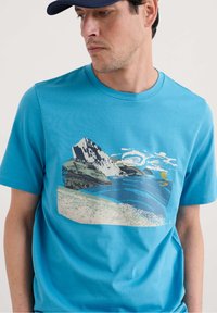 Turkio spalvos trumpų rankovių marškinėliai iš medvilnės, su kalno ir vandenyno grafika nuobodžiose spalvose, tekstūruotais bangų ir saulės piešiniais.