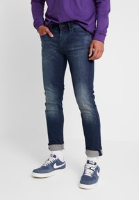 Slim-fit mörkblå denimjeans med lätt blekning, uppvikta byxben och bälteshål, ihop med blå och vita sneakers.