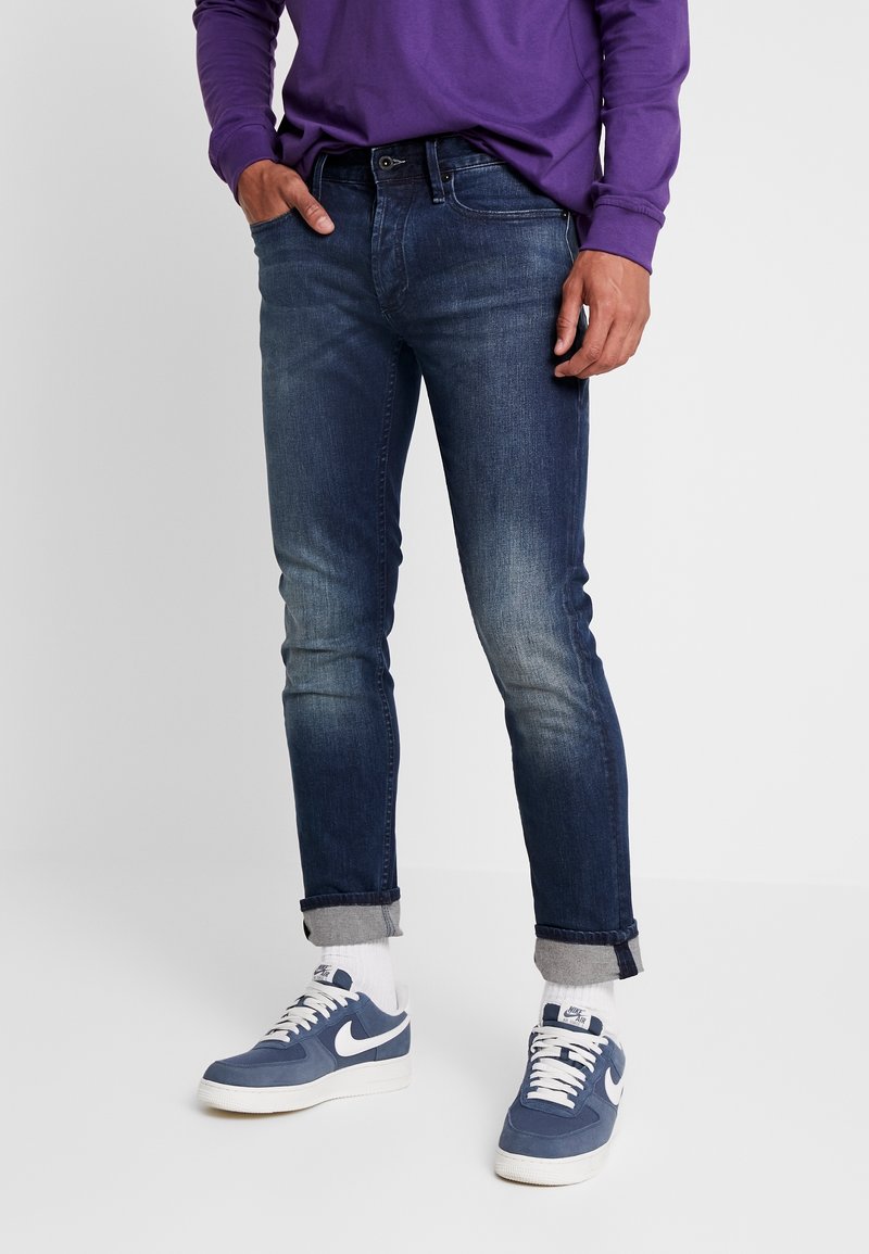 Slim-fit mörkblå denimjeans med lätt blekning, uppvikta byxben och bälteshål, ihop med blå och vita sneakers.