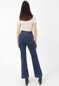 Vaqueros acampanados de tiro alto en denim azul oscuro, con dos bolsillos traseros y costuras visibles, combinados con una camiseta ajustada beige.