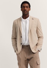 Beige Blazer mit schwarzen Knöpfen, taillierte Passform, eine Brusttasche, über einem weißen Button-Up-Hemd getragen, kombiniert mit beigen Cargo-Hosen.