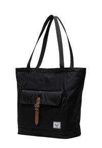 Sac en toile noir avec deux bretelles, une poche frontale fermée par une sangle en cuir marron et une étiquette de marque blanche sur le côté.