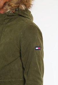 Olivgrön jacka med texturerad yta, försedd med en avtagbar fuskpälsmössa och en liten logotypapplikation på vänster ärm.