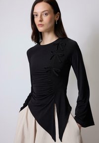 Maglietta nera a maniche lunghe con dettagli arricciati e tre fiocchi neri di tessuto sul lato sinistro; presenta un orlo curvo e asimmetrico.
