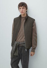 Beżowa kamizelka bez rękawów nałożona na brązowy, teksturowany sweter, zestawiona z ciemnoszarymi dżinsami. Model stoi na tle jasnego tła.